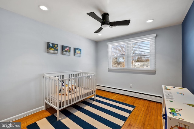 bedroom with a crib, a baseboard heating unit, ceiling fan, and wood-type flooring