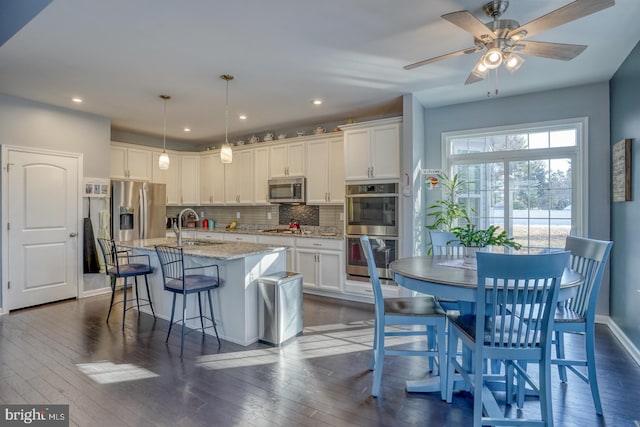 kitchen with a kitchen island with sink, stainless steel appliances, decorative backsplash, white cabinetry, and decorative light fixtures