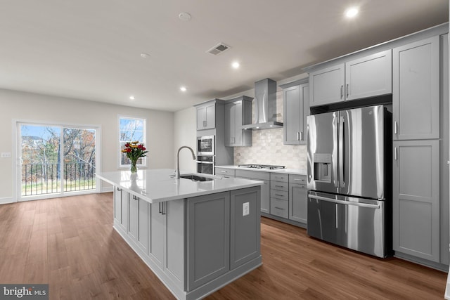 kitchen with wall chimney range hood, gray cabinets, a kitchen island with sink, appliances with stainless steel finishes, and sink