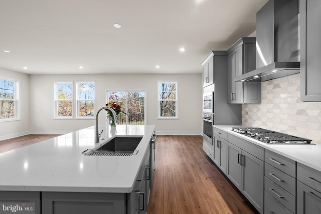 kitchen with sink, stainless steel appliances, gray cabinets, a center island with sink, and wall chimney range hood