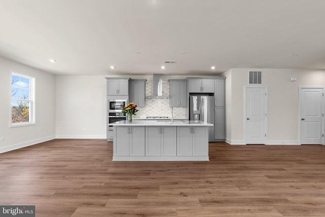 kitchen featuring stainless steel appliances, a center island with sink, gray cabinetry, and wall chimney exhaust hood