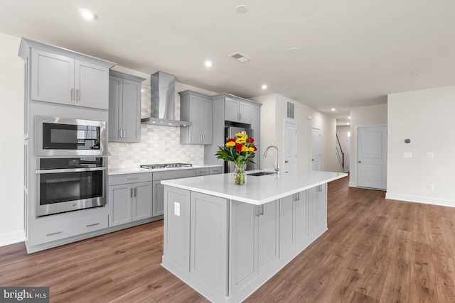 kitchen with an island with sink, appliances with stainless steel finishes, wall chimney exhaust hood, and gray cabinets