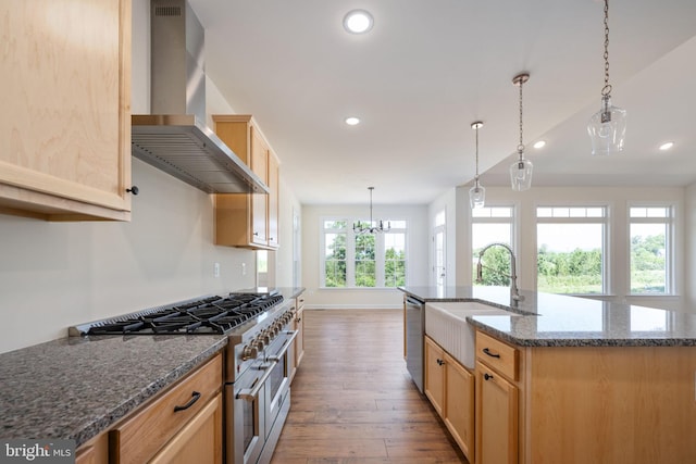 kitchen featuring wall chimney exhaust hood, stainless steel appliances, light brown cabinets, a kitchen island with sink, and sink