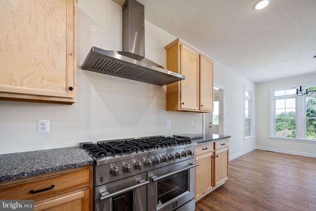 kitchen with dark stone counters, range with two ovens, wall chimney range hood, and light brown cabinets