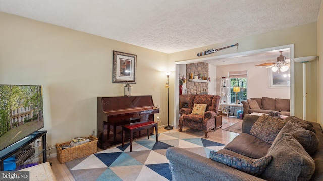 living room with ceiling fan, a stone fireplace, a textured ceiling, and light hardwood / wood-style flooring