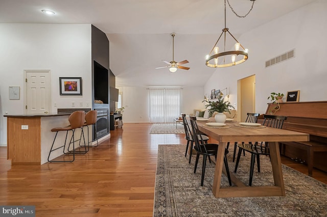 dining room with wood-type flooring, ceiling fan with notable chandelier, a fireplace, and high vaulted ceiling