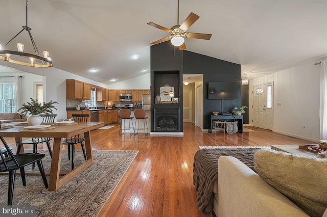 dining space featuring high vaulted ceiling, a large fireplace, ceiling fan with notable chandelier, and light wood-type flooring