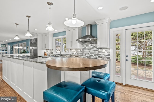 kitchen with built in fridge, white cabinets, wall chimney range hood, hanging light fixtures, and light wood-type flooring