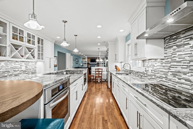 kitchen with pendant lighting, white cabinets, wall chimney exhaust hood, sink, and stainless steel oven