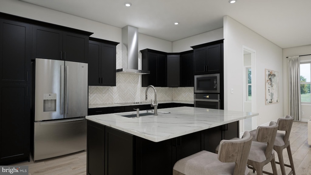 kitchen with light stone countertops, appliances with stainless steel finishes, wall chimney range hood, light hardwood / wood-style floors, and a kitchen island with sink