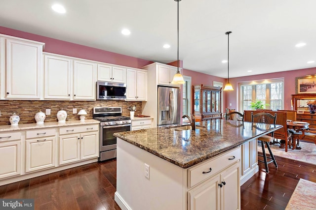 kitchen with dark stone countertops, appliances with stainless steel finishes, an island with sink, sink, and decorative light fixtures