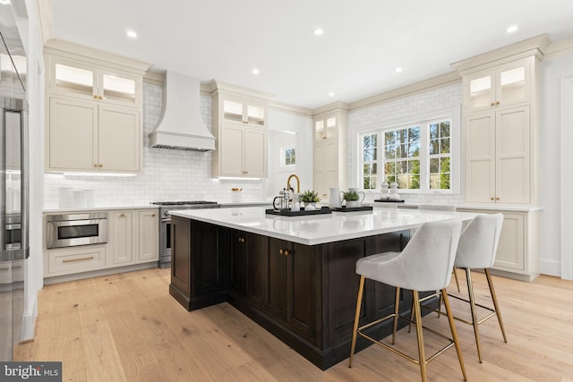 kitchen featuring custom exhaust hood, stainless steel appliances, an island with sink, light hardwood / wood-style floors, and white cabinetry