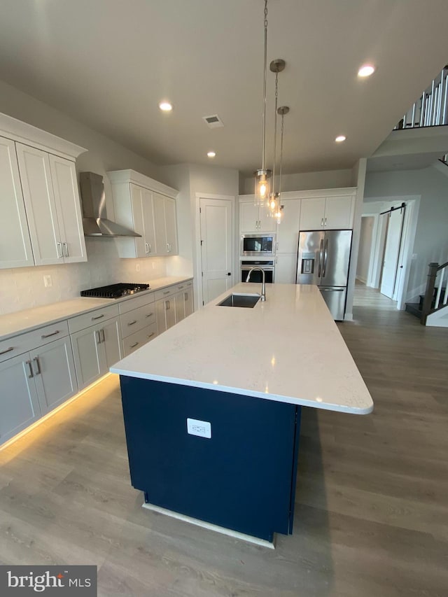 kitchen with white cabinets, appliances with stainless steel finishes, wall chimney range hood, and a kitchen island with sink