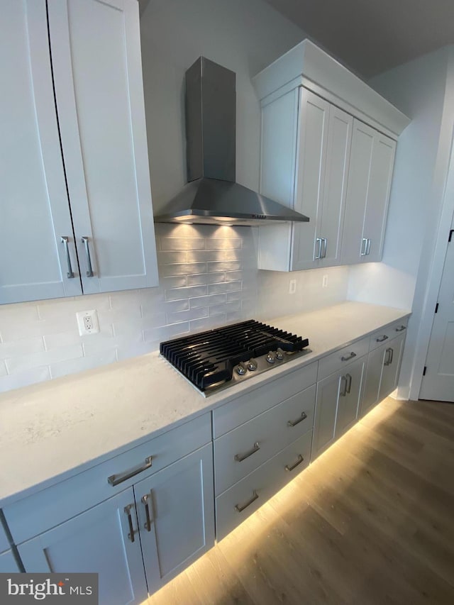 kitchen featuring wall chimney range hood, decorative backsplash, stainless steel gas cooktop, white cabinets, and wood-type flooring