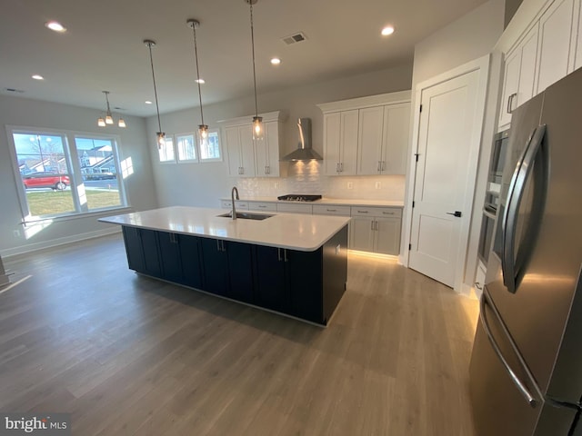 kitchen with decorative light fixtures, white cabinetry, stainless steel fridge, a center island with sink, and wall chimney range hood