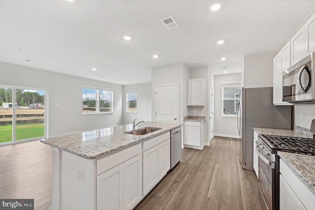 kitchen with stainless steel appliances, sink, white cabinetry, light stone counters, and an island with sink