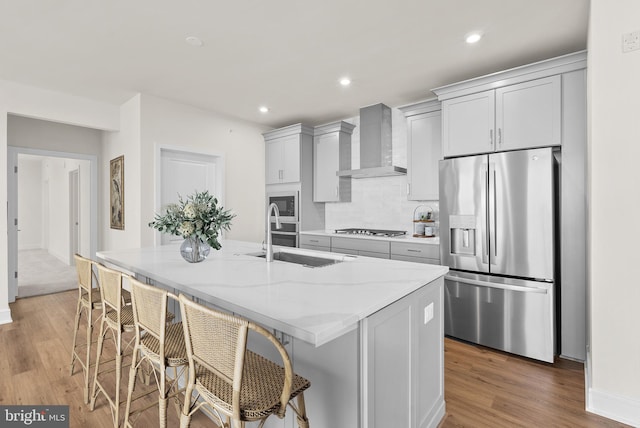 kitchen with stainless steel appliances, sink, wall chimney exhaust hood, an island with sink, and light stone countertops