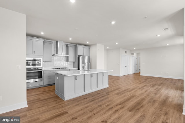 kitchen with a kitchen island with sink, gray cabinetry, stainless steel appliances, wall chimney exhaust hood, and backsplash