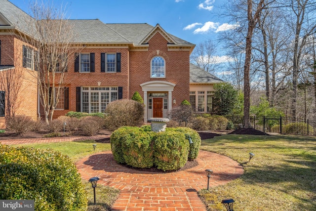 colonial-style house featuring a front yard, fence, brick siding, and roof with shingles