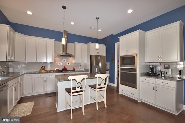 kitchen featuring wall chimney exhaust hood, hanging light fixtures, stainless steel appliances, a kitchen island, and dark stone countertops