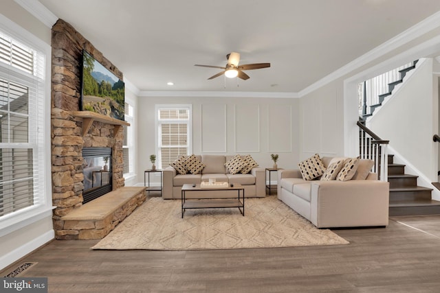 living room with light wood-type flooring, ceiling fan, ornamental molding, and a stone fireplace