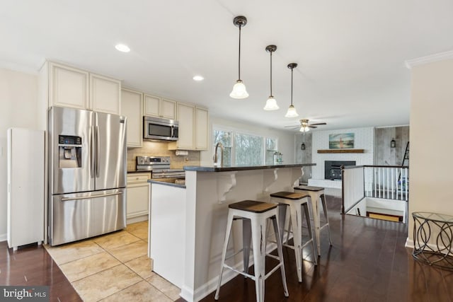 kitchen with cream cabinetry, a fireplace, dark countertops, appliances with stainless steel finishes, and a ceiling fan