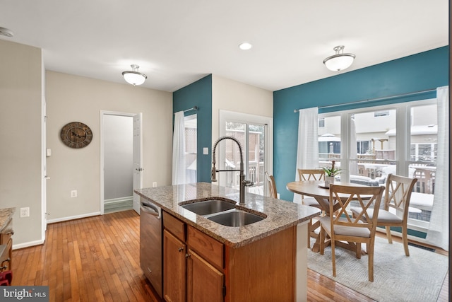 kitchen featuring dishwasher, sink, light wood-type flooring, light stone counters, and a center island with sink