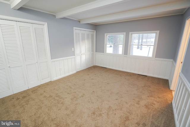 unfurnished bedroom with two closets, light carpet, ornamental molding, and beam ceiling