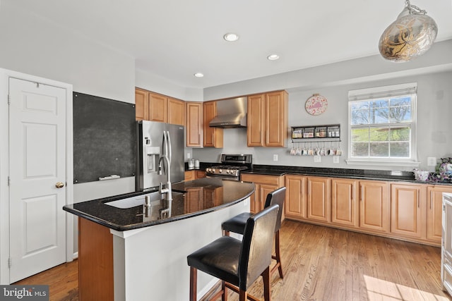 kitchen featuring wall chimney range hood, a center island with sink, sink, light hardwood / wood-style flooring, and appliances with stainless steel finishes