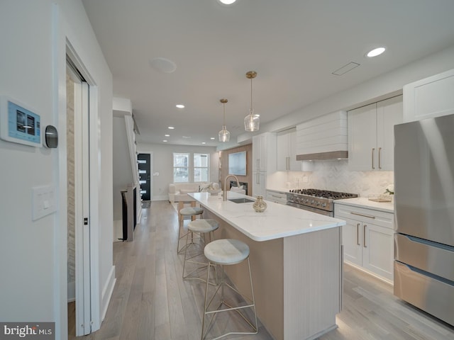 kitchen featuring premium range hood, appliances with stainless steel finishes, white cabinetry, hanging light fixtures, and a center island with sink