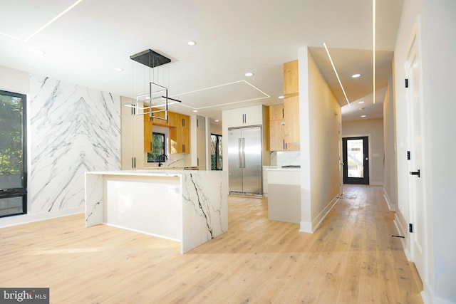 kitchen with stainless steel built in refrigerator, light brown cabinetry, hanging light fixtures, light wood-type flooring, and light stone countertops