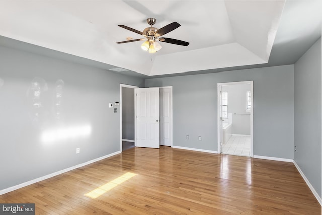 unfurnished bedroom with a tray ceiling, ensuite bath, ceiling fan, and light wood-type flooring