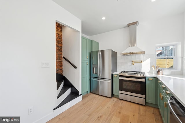 kitchen with green cabinets, stainless steel appliances, backsplash, light wood-type flooring, and wall chimney range hood