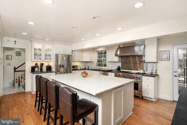 kitchen featuring backsplash, appliances with stainless steel finishes, light wood-style floors, a kitchen island, and wall chimney exhaust hood