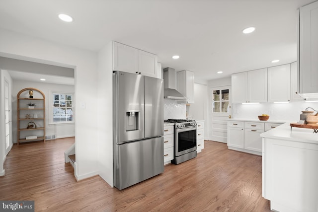 kitchen featuring wall chimney exhaust hood, stainless steel appliances, light hardwood / wood-style floors, backsplash, and white cabinets