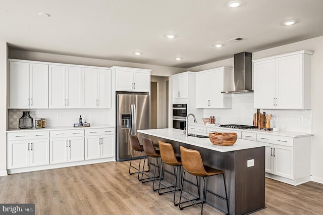 kitchen with wall chimney exhaust hood, stainless steel appliances, an island with sink, white cabinetry, and sink