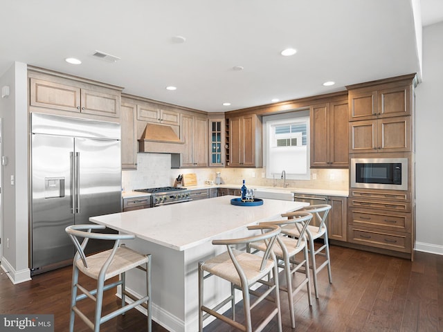 kitchen with a center island, glass insert cabinets, built in appliances, premium range hood, and a kitchen bar