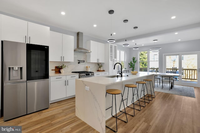 kitchen with decorative light fixtures, white cabinetry, an island with sink, wall chimney range hood, and appliances with stainless steel finishes