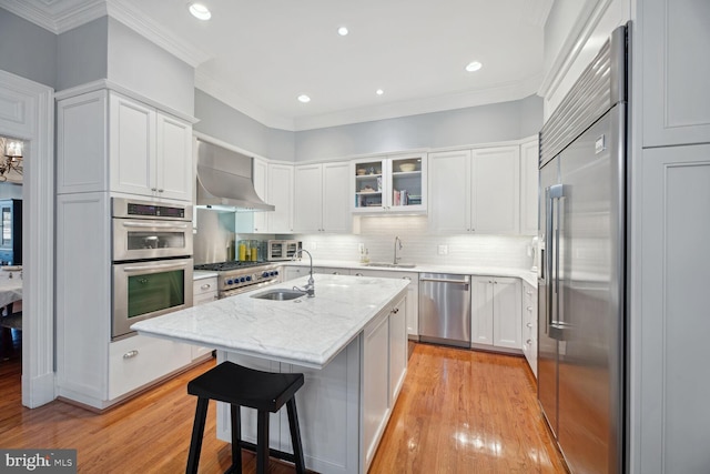 kitchen with appliances with stainless steel finishes, white cabinets, a kitchen island with sink, light stone countertops, and wall chimney range hood