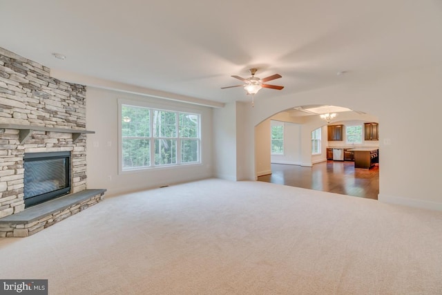 unfurnished living room with ceiling fan with notable chandelier, a stone fireplace, and dark colored carpet