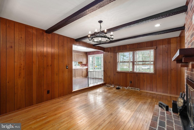 unfurnished living room featuring beam ceiling, a brick fireplace, wood walls, and a notable chandelier