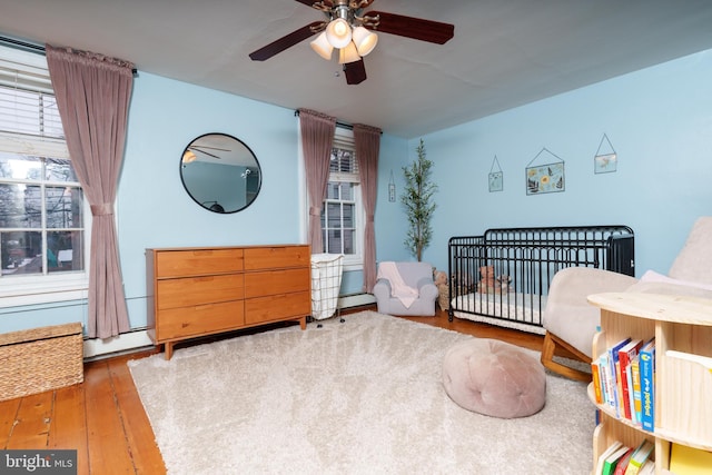 bedroom featuring ceiling fan, hardwood / wood-style floors, a baseboard radiator, and a crib