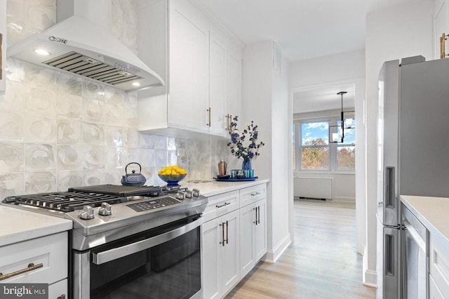 kitchen with appliances with stainless steel finishes, decorative backsplash, white cabinets, wall chimney exhaust hood, and light hardwood / wood-style flooring