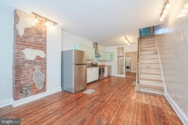 kitchen with appliances with stainless steel finishes, wall chimney exhaust hood, washer / clothes dryer, green cabinetry, and light wood-type flooring