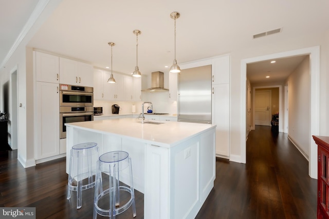 kitchen featuring an island with sink, appliances with stainless steel finishes, hanging light fixtures, wall chimney exhaust hood, and white cabinets