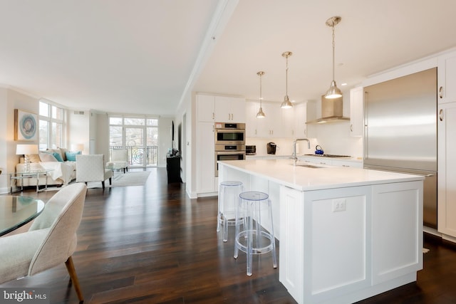 kitchen with appliances with stainless steel finishes, wall chimney exhaust hood, white cabinetry, hanging light fixtures, and a kitchen island with sink