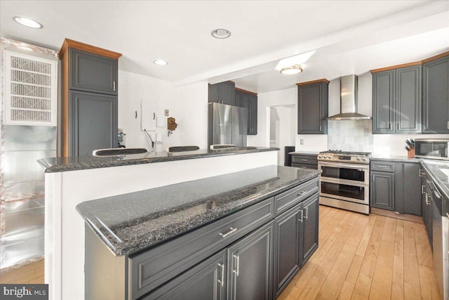 kitchen with appliances with stainless steel finishes, wall chimney exhaust hood, dark stone counters, and a kitchen island with sink