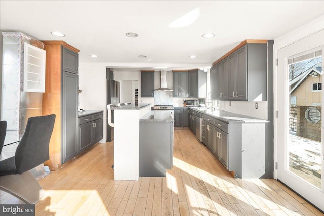 kitchen with appliances with stainless steel finishes, light stone countertops, a kitchen island, light hardwood / wood-style flooring, and wall chimney range hood