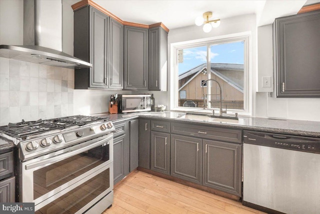 kitchen featuring appliances with stainless steel finishes, wall chimney exhaust hood, dark stone counters, light hardwood / wood-style floors, and sink