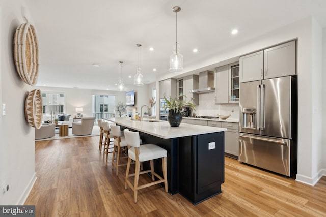 kitchen with wall chimney range hood, gray cabinetry, stainless steel appliances, an island with sink, and decorative light fixtures